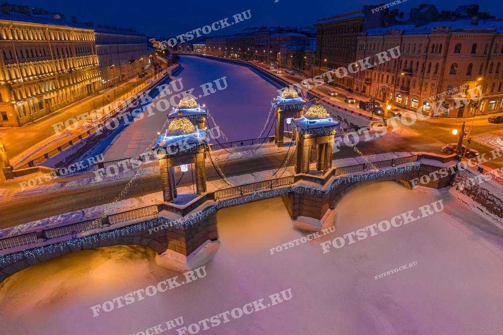 На фотографии изображен мост Ломоносова в Санкт-Петербурге, украшенный рождественскими гирляндами. Мост перекинут через замерзшую реку Фонтанку, создавая атмосферу зимнего праздника. Окружающие здания подсвечены теплым светом уличных фонарей, добавляя уют и красоту ночному зимнему Петербургу.